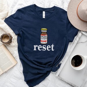 Peut inclure: T-shirt bleu marine avec un dessin d'un flacon de m&eacute;dicament et le texte "HALDOL HALOPERIDOL INJECTION USP", ainsi que le mot "reset" en blanc. Le t-shirt est pos&eacute; sur une surface blanche avec une tasse de caf&eacute; et un chapeau.