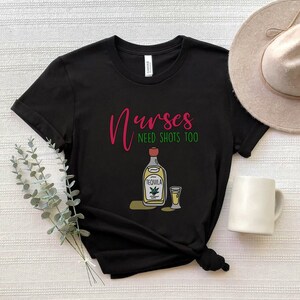 May include: Black t-shirt with the text "Nurses need shots too" in pink and green. A cartoon image of a bottle of tequila and a shot glass are below the text.