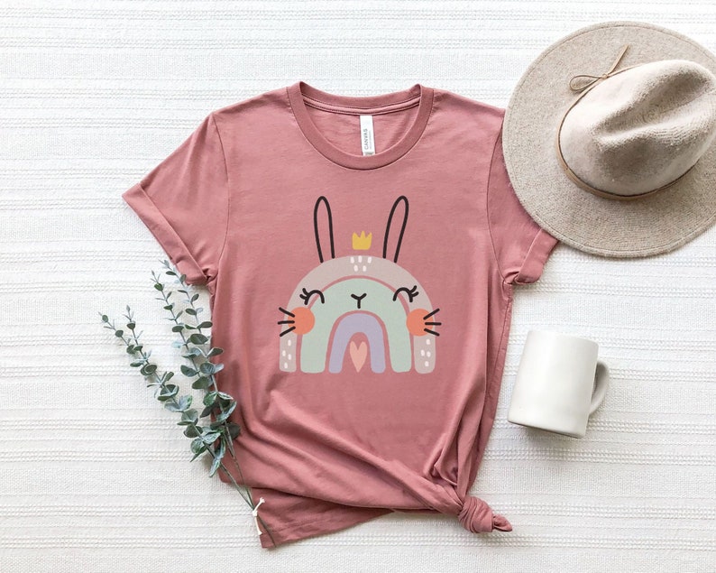 Może przedstawiać: R&oacute;żowy T-shirt z grafiką przedstawiającą kr&oacute;lika w koronie siedzącego na tęczy. Kr&oacute;lik ma sercowaty nos i zamknięte oczy.