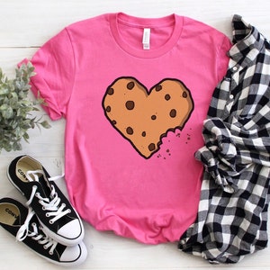 May include: Pink t-shirt with a brown chocolate chip cookie shaped like a heart with a bite taken out of it.