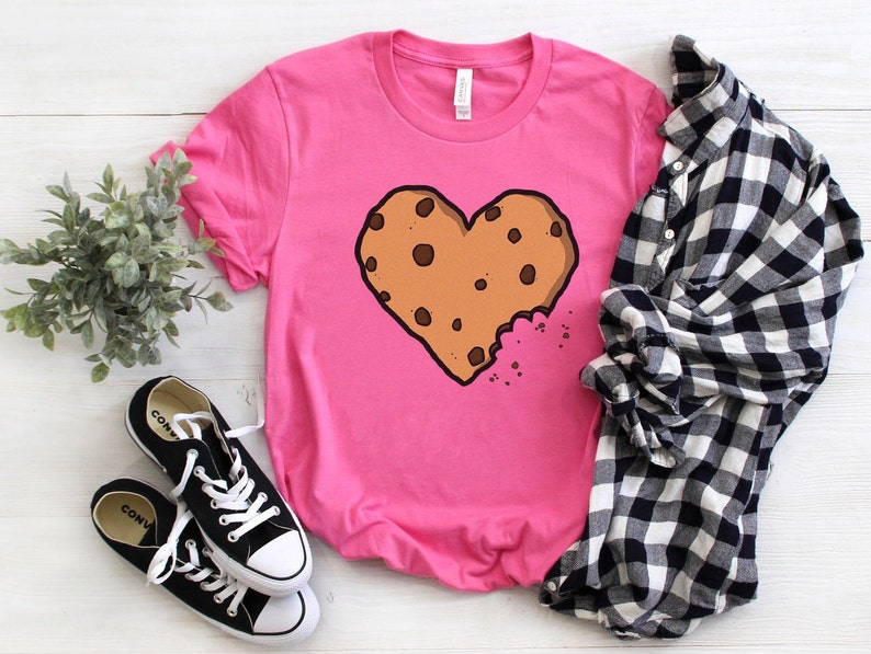 May include: Pink t-shirt with a brown chocolate chip cookie shaped like a heart with a bite taken out of it.