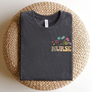May include: Dark gray t-shirt with a colorful floral design and the word "NURSE" in a retro font. The flowers include shades of blue, pink, yellow, and green. The shirt is folded neatly on a woven, circular surface.