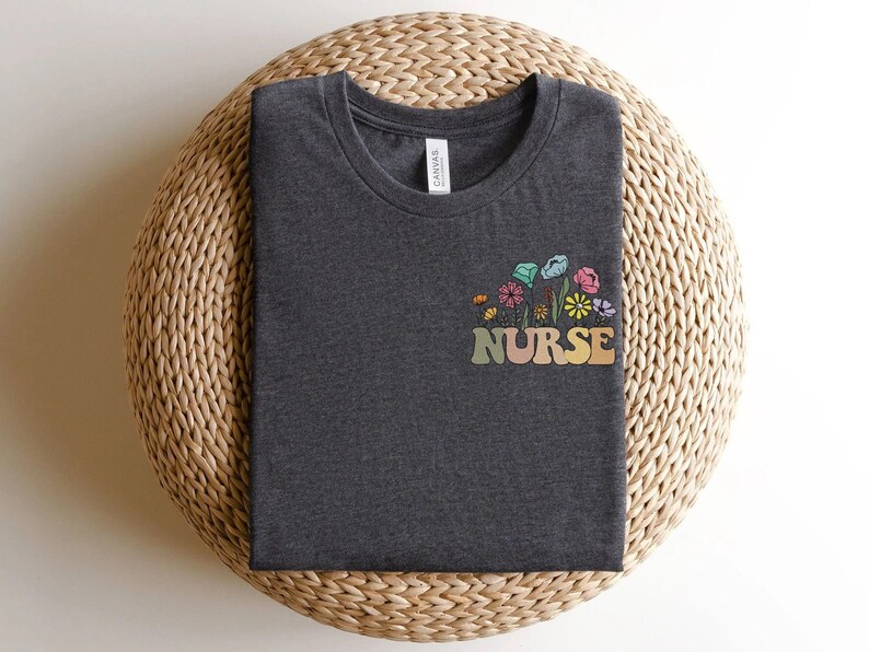 May include: Dark gray t-shirt with a colorful floral design and the word "NURSE" in a retro font. The flowers include shades of blue, pink, yellow, and green. The shirt is folded neatly on a woven, circular surface.