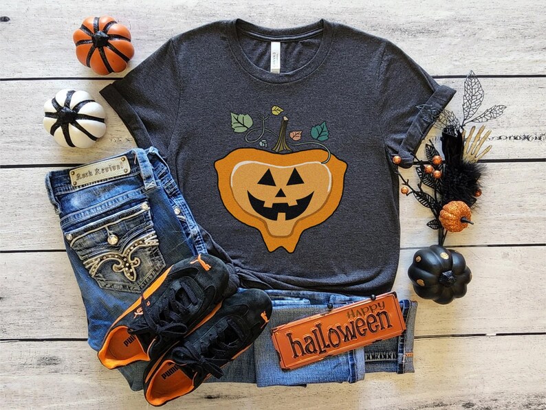 May include: A dark grey t-shirt with a graphic of a melting jack-o'-lantern with a happy face. The shirt is surrounded by jeans, sneakers, and Halloween decorations.