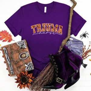 Puede incluir: Camiseta morada con el texto "Trauma Nurse" en naranja con tem&aacute;tica de Halloween. La camiseta se muestra sobre una superficie blanca con una escoba, un libro y una ara&ntilde;a.