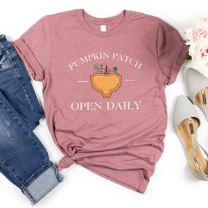 K&ouml;nnte beinhalten: Ein mauvefarbenes T-Shirt mit dem Text "Pumpkin Patch Open Daily" und einer stilisierten K&uuml;rbisgrafik.