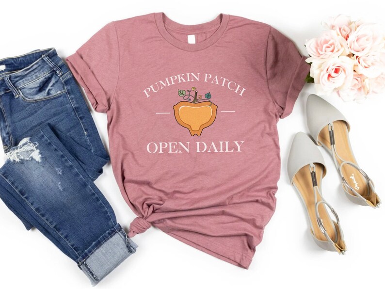 K&ouml;nnte beinhalten: Ein mauvefarbenes T-Shirt mit dem Text "Pumpkin Patch Open Daily" und einer stilisierten K&uuml;rbisgrafik.