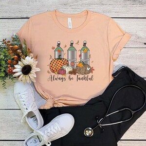 May include: A peach-colored t-shirt with a graphic of three green oxygen tanks, pumpkins, and fall leaves. The text "Always be Tankful" is printed below the graphic.