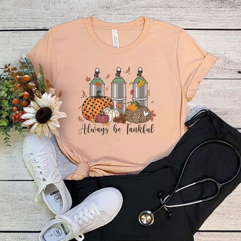 May include: A peach-colored t-shirt with a graphic of three green oxygen tanks, pumpkins, and fall leaves. The text "Always be Tankful" is printed below the graphic.