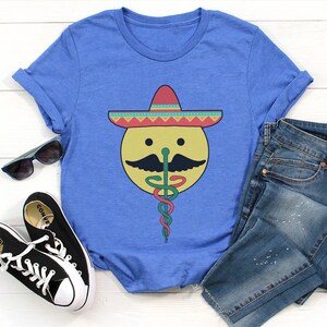 K&ouml;nnte beinhalten: Ein blaues T-Shirt mit einem Aufdruck eines l&auml;chelnden gelben Gesichts, das einen bunten Sombrero und einen gr&uuml;n-roten medizinischen Caduceus tr&auml;gt.
