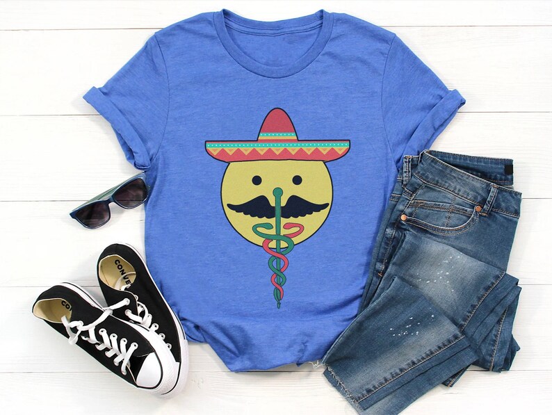K&ouml;nnte beinhalten: Ein blaues T-Shirt mit einem Aufdruck eines l&auml;chelnden gelben Gesichts, das einen bunten Sombrero und einen gr&uuml;n-roten medizinischen Caduceus tr&auml;gt.
