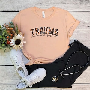 Puede incluir: Una camiseta de color melocot&oacute;n con un gr&aacute;fico negro y naranja que dice "Trauma Nurse" en una fuente universitaria. El gr&aacute;fico presenta un patr&oacute;n de calabazas y fantasmas.