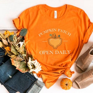 K&ouml;nnte beinhalten: Orangefarbenes T-Shirt mit dem Text "Pumpkin Patch Open Daily" und einer Grafik einer K&uuml;rbis mit Stiel und Bl&auml;ttern. Das Shirt liegt auf einer wei&szlig;en Holzoberfl&auml;che mit einem Paar braunen Sandalen, einem B&uuml;ndel Herbstlaub und einem kleinen orangefarbenen K&uuml;rbis.