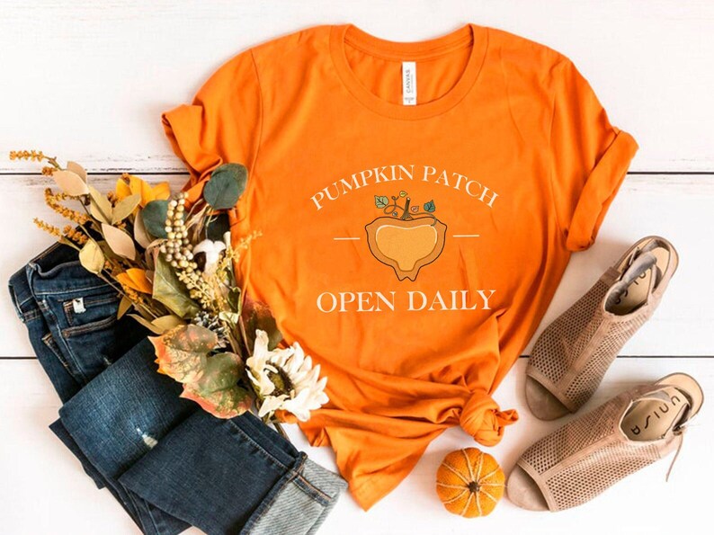 K&ouml;nnte beinhalten: Orangefarbenes T-Shirt mit dem Text "Pumpkin Patch Open Daily" und einer Grafik einer K&uuml;rbis mit Stiel und Bl&auml;ttern. Das Shirt liegt auf einer wei&szlig;en Holzoberfl&auml;che mit einem Paar braunen Sandalen, einem B&uuml;ndel Herbstlaub und einem kleinen orangefarbenen K&uuml;rbis.