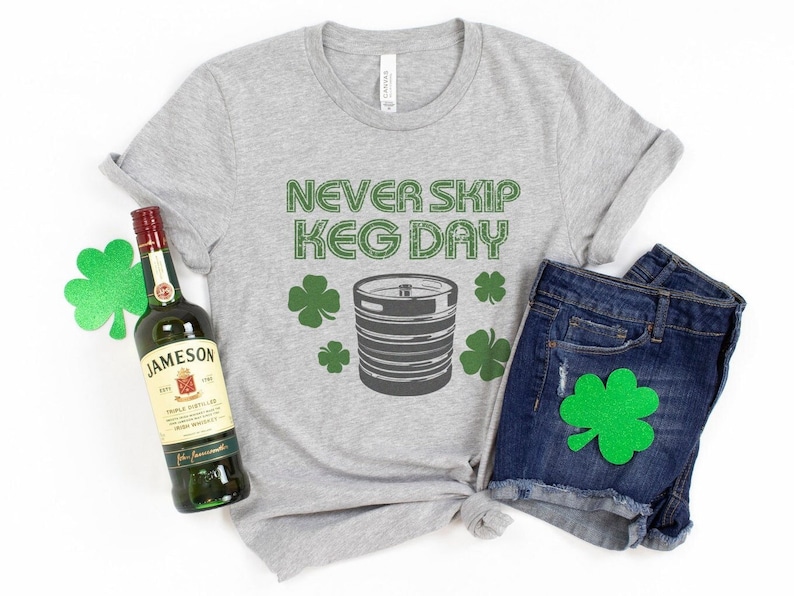 May include: A gray t-shirt with the text "Never Skip Keg Day" and a green shamrock design. A bottle of Jameson Irish Whiskey and a pair of blue denim shorts with a green shamrock patch are also pictured.