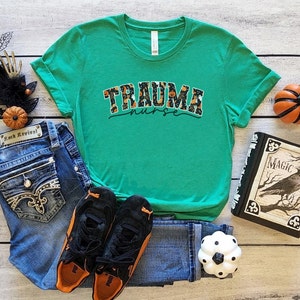 Puede incluir: Una camiseta verde con un gr&aacute;fico de tem&aacute;tica de Halloween que dice "Trauma Nurse". El gr&aacute;fico presenta calabazas, murci&eacute;lagos y fantasmas.