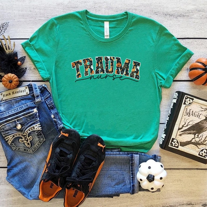 Puede incluir: Una camiseta verde con un gr&aacute;fico de tem&aacute;tica de Halloween que dice "Trauma Nurse". El gr&aacute;fico presenta calabazas, murci&eacute;lagos y fantasmas.