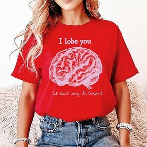 May include: A red t-shirt featuring the words "I lobe you" and a pink brain graphic. Beneath the brain, the text reads "but don't worry, it's temporal". The top is a short-sleeved crew neck.