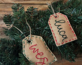 May include: Two beige felt gift tags with stitched edges and twine hangers rest on a bed of green pine needles. One tag reads "Wes" in red thread, the other reads "Luca" in black thread. The tags are likely for Christmas or other holiday gifts.