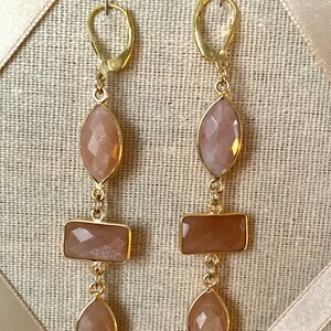 May include: A pair of gold dangle earrings with three peach-colored sunstone gemstones on each earring. The earrings have a hook closure.