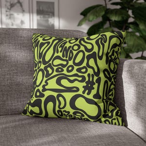 May include: Square throw pillow with a lime green background and a black abstract design. The design includes the text "LANDO" and the number "106". The pillow is resting on a gray sofa.