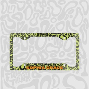 Green and black f1 themed lando norris car license plate frame is shown against a black and white lando norris pattern. Personalised name in orange is on the bottom edge of the frame. 

Lando Norris F1 License Plate Frame: Green and black pattern, Aluminum