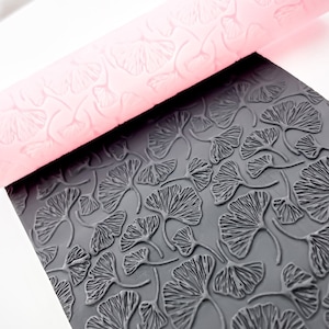 Op de afbeelding: Een roze deegroller en een donkergrijze, getextureerde plaat, beide met een ginkgo bladpatroon. De deegroller is cilindrisch en de plaat is rechthoekig. Het ginkgo bladontwerp is op beide items in reliëf aangebracht.