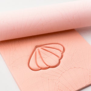 Könnte beinhalten: Eine rosa Silikon-Nudelholz mit einem geprägten Muschel-Design auf der Oberfläche. Das Nudelholz wird auf einer ebenen Fläche ausgerollt.
