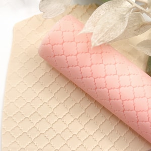 Könnte beinhalten: Ein rosafarbener Silikon-Nudelholz mit einem sich wiederholenden geometrischen Muster. Das Nudelholz liegt auf einer beigefarbenen Silikonmatte mit dem gleichen Muster.