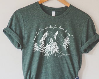 May include: A green t-shirt with a white graphic of mountains and trees. The text on the shirt reads "come to the woods for here is real".