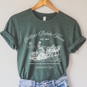Puede incluir: Una camiseta verde con un gráfico blanco de un granjero en un arado tirado por caballos. El gráfico dice "Collins' Potato Farm Est. 1813" y "Perfecto para excelentes patatas hervidas Hunsford, Kent".