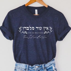 May include: A navy blue t-shirt with rolled-up sleeves, paired with denim shorts, hangs on a wooden hanger. The shirt features white text in Hebrew and English: "EIN OD MILVADO, There Is None But Him."
