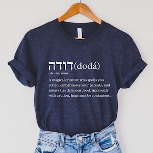Peut inclure: Un t-shirt bleu marine avec du texte blanc qui dit "דודה (doda) [do-da] noun A magical creature who spoils you rotten, embarrasses your parents, and always has delicious food. Approach with caution, hugs may be contagious."