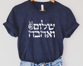 May include: Dark blue t-shirt with white Hebrew text and a peace sign. The text appears to read "Shalom Ve'ahava", which translates to "Peace and Love". The shirt is paired with blue denim shorts.