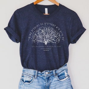 Könnte beinhalten: Ein marineblaues T-Shirt mit einem weißen Baumdesign und hebräischem Text. Der Text lautet "Es gibt einen Baum des Lebens für diejenigen, die sich daran klammern, und diejenigen, die sich daran klammern, sind gesegnet." Sprichwörter 3:18.