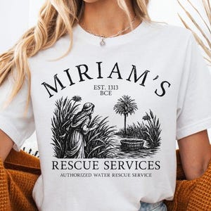 Pode incluir: Camiseta branca com um gráfico preto. O gráfico inclui o texto "Miriam's Rescue Services" e uma ilustração de uma pessoa perto da água e plantas. A camiseta também apresenta "Est. 1313 a.C." e "Authorized Water Rescue Service".