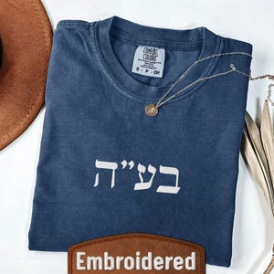 Può includere: Viene mostrata una maglietta blu navy con testo ebraico bianco. Una collana d'oro con un ciondolo poggia sulla maglietta. Un cappello marrone e foglie secche sono sullo sfondo. La maglietta ha una piccola etichetta che dice "Comfort Colors". Una toppa in pelle marrone in basso dice "Embroidered".