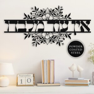 May include: Black metal wall art with the Hebrew word "Shalom" in a decorative font, surrounded by floral designs. The art is mounted on a white wall with a calendar, books, and a vase of flowers in the foreground. The text "Powder Coated Steel" is visible in a circle below the art.