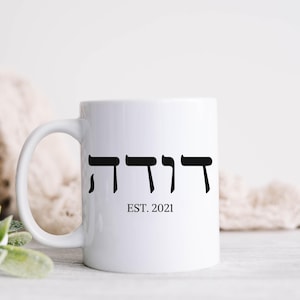 May include: White ceramic coffee mug with a black Hebrew word and the text "EST. 2021" printed on it.