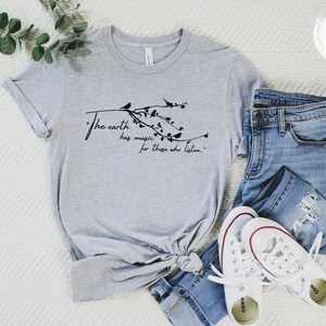 May include: A gray t-shirt with a black graphic that reads "The earth has music for those who listen." The graphic features a branch with birds and leaves. The t-shirt is laid out on a white surface with a pair of white sneakers and blue jeans.