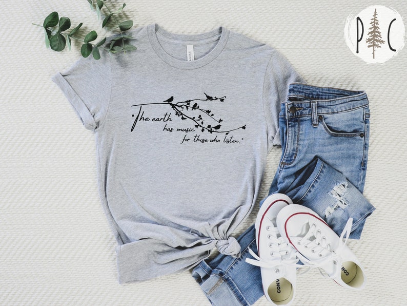 May include: A gray t-shirt with a black graphic that reads "The earth has music for those who listen." The graphic features a branch with birds and leaves. The t-shirt is laid out on a white surface with a pair of white sneakers and blue jeans.