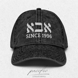 May include: A black, distressed baseball cap with white embroidered text. The text includes Hebrew characters above the words "SINCE 1996". The cap has a curved brim and a fabric strap closure. The bottom of the image has the word "pacific" in cursive.