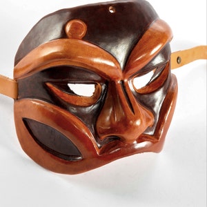 Puede incluir: Una m&aacute;scara de cuero marr&oacute;n con una cara estilizada, que presenta una nariz grande y ojos prominentes. La m&aacute;scara tiene una correa de cuero marr&oacute;n.
