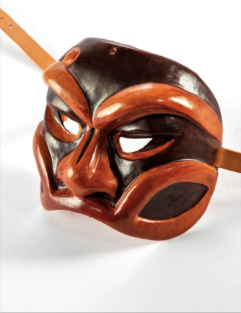 Puede incluir: Una m&aacute;scara de cuero marr&oacute;n y negro con una expresi&oacute;n dram&aacute;tica. La m&aacute;scara tiene una nariz grande y prominente y cejas exageradas. La m&aacute;scara est&aacute; dise&ntilde;ada con una correa para llevarla.