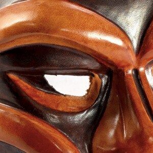 Puede incluir: Primer plano de una m&aacute;scara de cuero marr&oacute;n con un dise&ntilde;o negro. La m&aacute;scara tiene un gran agujero redondo para el ojo y un peque&ntilde;o agujero redondo en la frente.