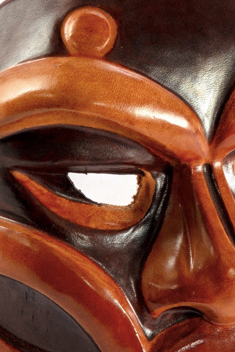 Puede incluir: Primer plano de una m&aacute;scara de cuero marr&oacute;n con un dise&ntilde;o negro. La m&aacute;scara tiene un gran agujero redondo para el ojo y un peque&ntilde;o agujero redondo en la frente.