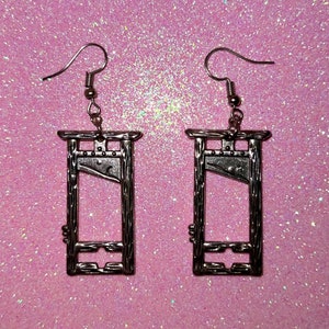 May include: A pair of silver earrings shaped like guillotines. The earrings have a detailed design and are hanging from silver hooks.