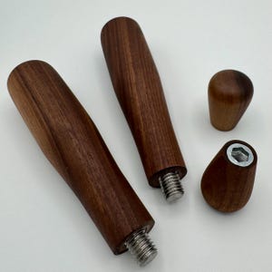 Könnte beinhalten: Vier Holzmöbelbeschläge. Zwei lange, zylindrische Griffe mit Gewindeenden aus Metall, ein kleiner runder Knopf und ein konisches Teil mit einer Sechskantmutter. Alle Teile sind aus dunkelbraunem Holz gefertigt.