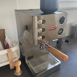 Könnte beinhalten: Espressomaschine aus Edelstahl mit Holzgriff und schwarzer Tasse. Die Maschine trägt den Markennamen "LELIT". Ein Kaffeestampfer mit Holzgriff steht auf der Theke neben der Maschine.