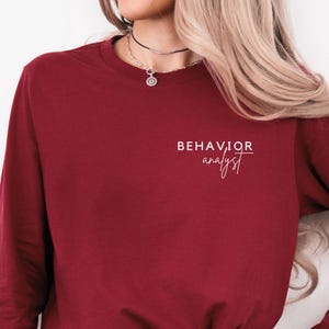 May include: A long-sleeved cardinal red shirt with the words "BEHAVIOR analyst" printed in white. The shirt is a solid color and has a crew neck. The text is centered on the chest.